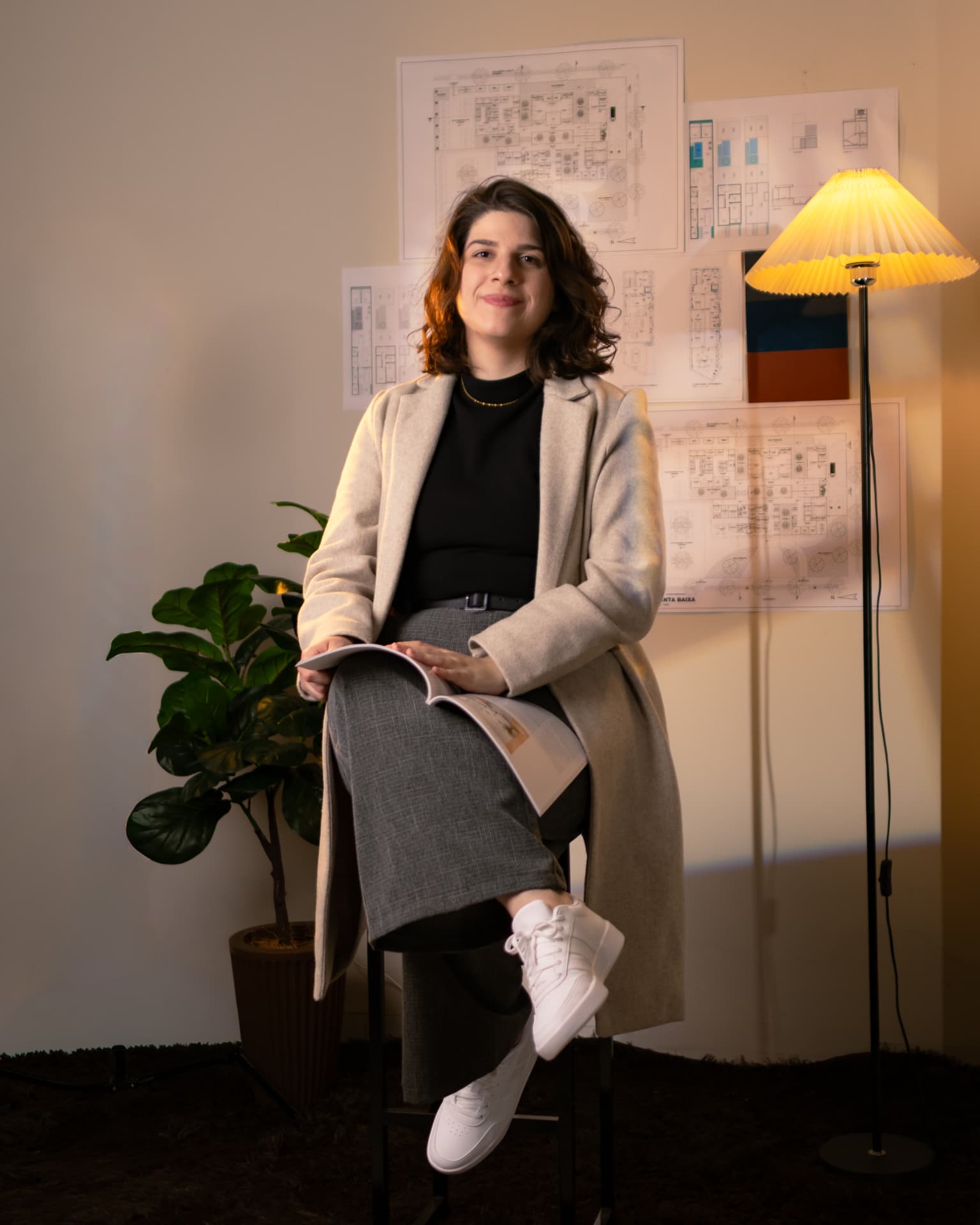 Luana Obici, arquiteta, sentada com projeto no colo e desenhos arquitetônicos ao fundo